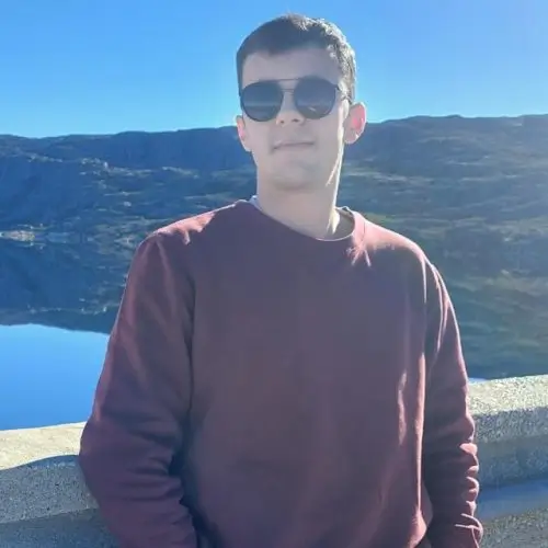 André, Projektleiter bei AkiByte, mit Sonnenbrille und bordeauxrotem Pullover an einem malerischen Bergsee