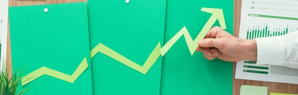 Hand hält Papierpfeil, der aufsteigend auf grünen Diagrammen zeigt und Geschäftswachstum durch SEO-Investitionen symbolisiert