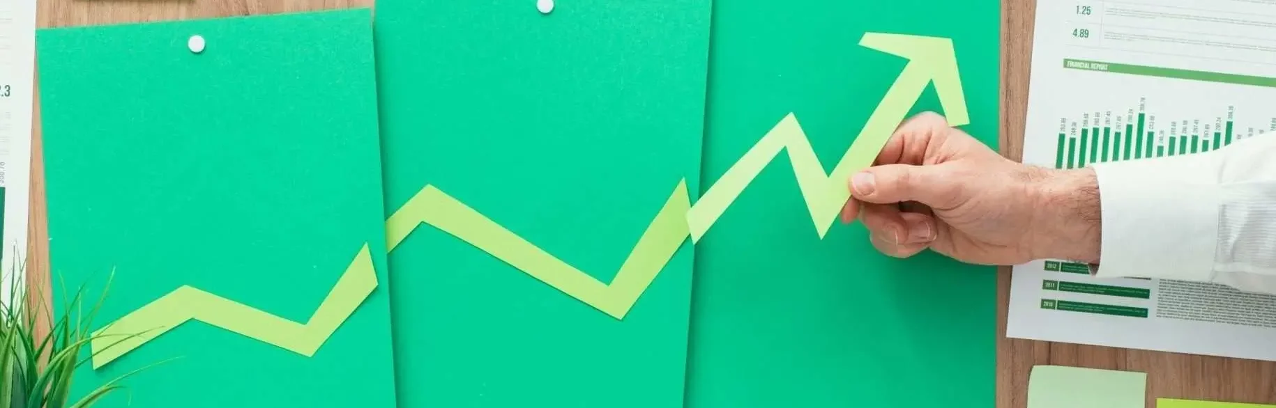 Hand hält Papierpfeil, der aufsteigend auf grünen Diagrammen zeigt und Geschäftswachstum durch SEO-Investitionen symbolisiert